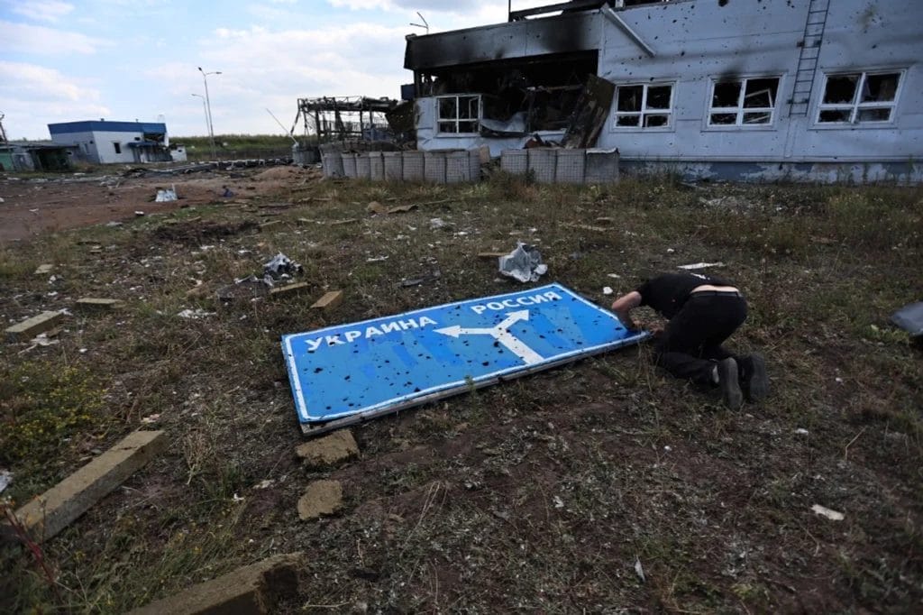 Homem examinando uma placa de sinalização danificada, uma indicando a direção da Ucrânia e a outra, da Rússia.